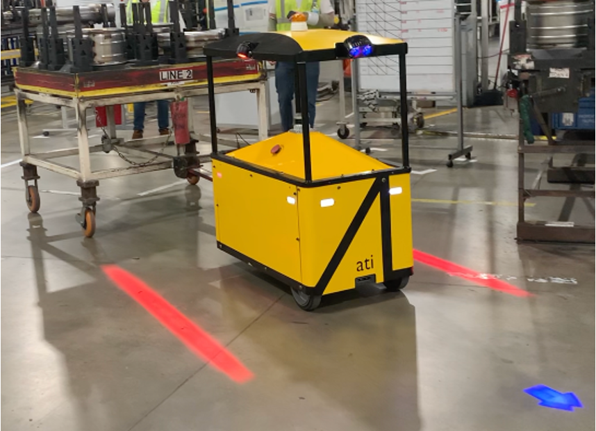 Sherpa tug-bot shining red laser lights on a concrete floor to left and right. A blue laser arrow is shown in front. Sherpa tows a rolling rack carrying car parts. An engineer stands in the background with a PS4 controller.