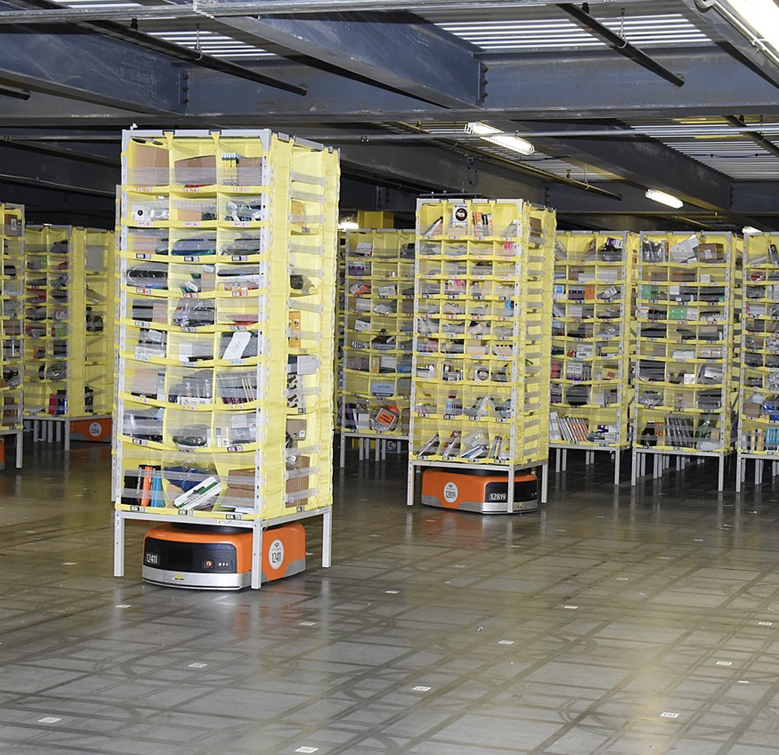squat orange Kiva robots in a dimly lit low-ceiling warehouse. Each robot carries a stack of yellow trays. Cubbies in the trays hold Amazon products. There are grid marks on the concrete floor and white&black QR codes every 4 feet on the gridlines.
